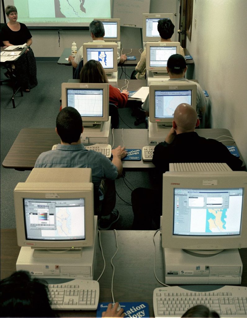 Adults follow a computer training session seated at desks in a bright classroom.